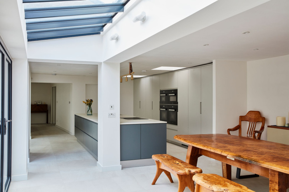 This is an example of a large contemporary single-wall kitchen in London with recessed-panel cabinets, grey cabinets, black appliances, an island, multi-coloured floors and white worktops.