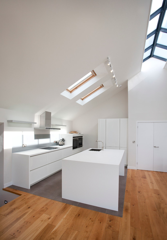 Inspiration for a medium sized contemporary single-wall open plan kitchen in Cornwall with a single-bowl sink, flat-panel cabinets, white cabinets, composite countertops, white splashback, ceramic splashback, stainless steel appliances, slate flooring, an island, grey floors, white worktops and a vaulted ceiling.