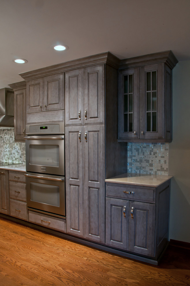 Mid-sized transitional l-shaped medium tone wood floor eat-in kitchen photo in Chicago with raised-panel cabinets, gray cabinets, quartz countertops, gray backsplash, mosaic tile backsplash, stainless steel appliances, an island and an undermount sink