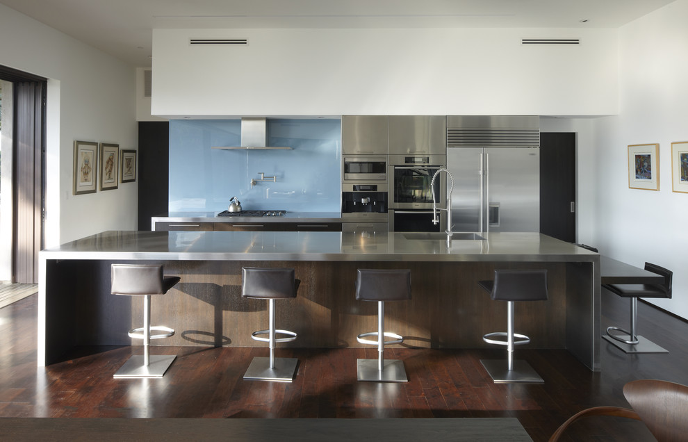Medium sized modern galley kitchen/diner in Los Angeles with an integrated sink, stainless steel worktops, blue splashback, glass sheet splashback, flat-panel cabinets, stainless steel appliances, an island, dark hardwood flooring and stainless steel cabinets.