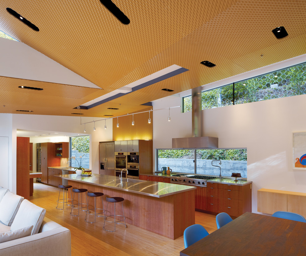 Идея дизайна: кухня-гостиная среднего размера в стиле модернизм с техникой из нержавеющей стали, плоскими фасадами, фасадами цвета дерева среднего тона, столешницей из нержавеющей стали, монолитной мойкой и светлым паркетным полом