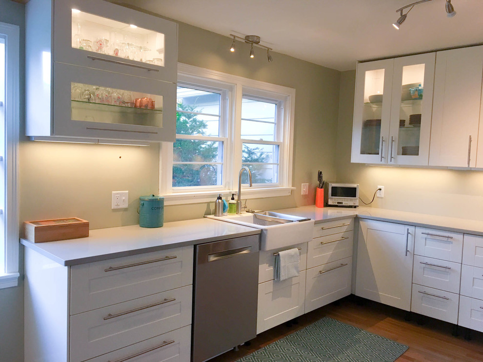 Medium sized retro l-shaped kitchen/diner in New York with a belfast sink, shaker cabinets, white cabinets, engineered stone countertops, stainless steel appliances, medium hardwood flooring, no island, brown floors and grey worktops.