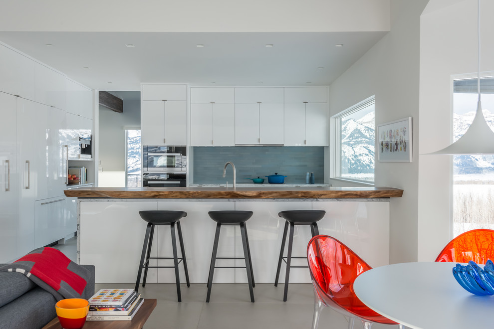 Свежая идея для дизайна: кухня-гостиная в современном стиле с плоскими фасадами, белыми фасадами, серым фартуком, полуостровом и окном - отличное фото интерьера