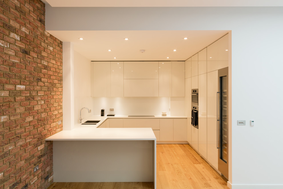 Example of a mid-sized trendy u-shaped light wood floor and brown floor kitchen design in London with a double-bowl sink, flat-panel cabinets, white cabinets, white backsplash, paneled appliances and solid surface countertops