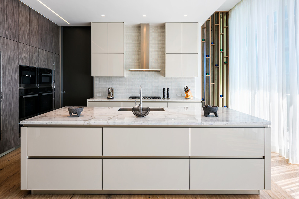 Contemporary l-shaped kitchen in Miami with a submerged sink, flat-panel cabinets, white cabinets, white splashback, mosaic tiled splashback, black appliances, medium hardwood flooring, an island, brown floors and white worktops.
