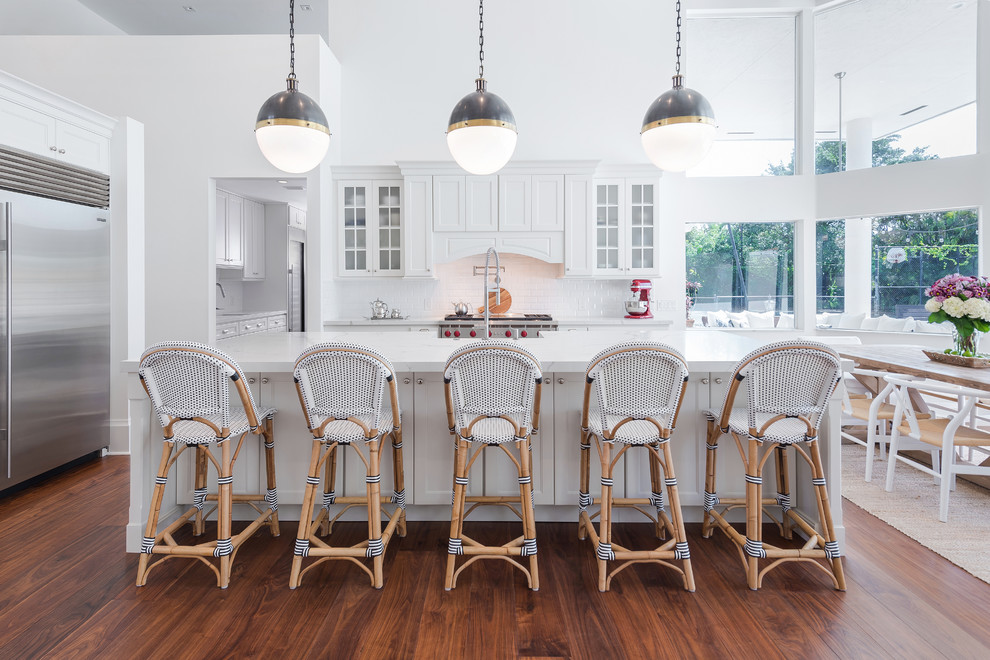 This is an example of a traditional l-shaped kitchen in Miami with a belfast sink, shaker cabinets, white cabinets, white splashback, stainless steel appliances, dark hardwood flooring, an island and brown floors.
