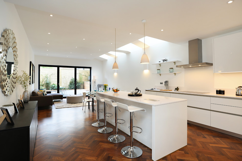 Expansive contemporary galley kitchen/diner in London with a built-in sink, flat-panel cabinets, white cabinets, engineered stone countertops, integrated appliances, dark hardwood flooring, brown floors, white splashback and an island.