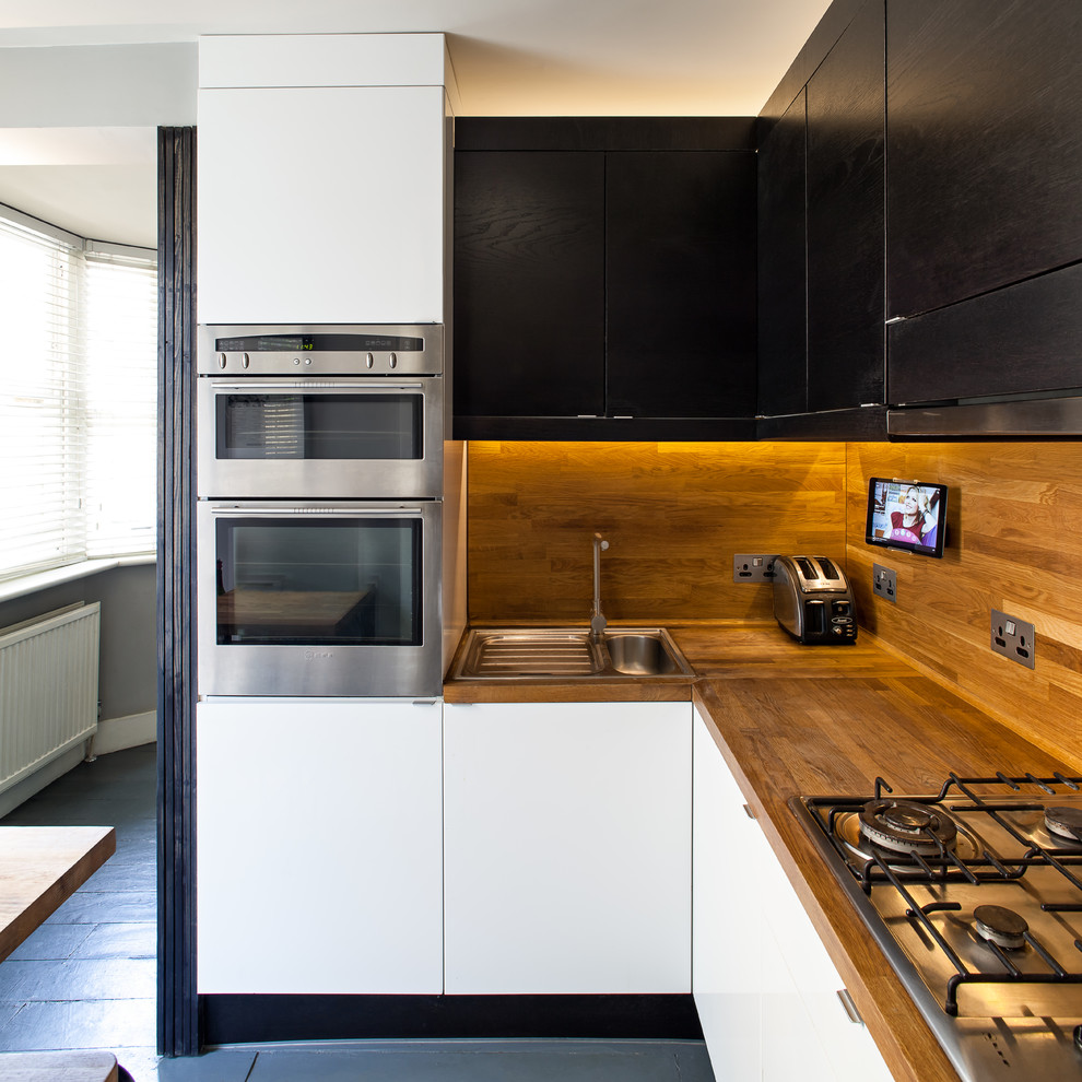 ロンドンにある低価格の小さなトランジショナルスタイルのおしゃれなキッチン (ドロップインシンク、フラットパネル扉のキャビネット、黒いキャビネット、木材カウンター、茶色いキッチンパネル、木材のキッチンパネル、シルバーの調理設備、塗装フローリング、グレーの床、茶色いキッチンカウンター) の写真
