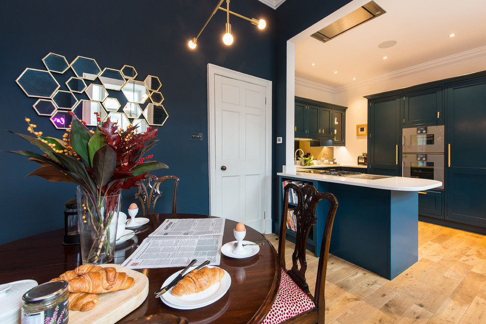 Example of a mid-sized minimalist u-shaped light wood floor eat-in kitchen design in Edinburgh with a drop-in sink, shaker cabinets, blue cabinets, solid surface countertops, mirror backsplash, stainless steel appliances, a peninsula and white countertops