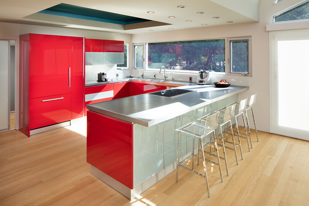 Mid-sized trendy u-shaped light wood floor and brown floor eat-in kitchen photo in San Francisco with a single-bowl sink, flat-panel cabinets, red cabinets, stainless steel countertops, white backsplash, glass sheet backsplash, stainless steel appliances, a peninsula and gray countertops