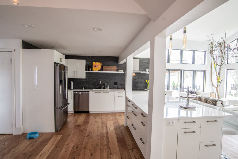 Example of a large trendy l-shaped medium tone wood floor and brown floor open concept kitchen design in New York with a single-bowl sink, flat-panel cabinets, white cabinets, quartz countertops, black backsplash, stone slab backsplash, stainless steel appliances, an island and white countertops