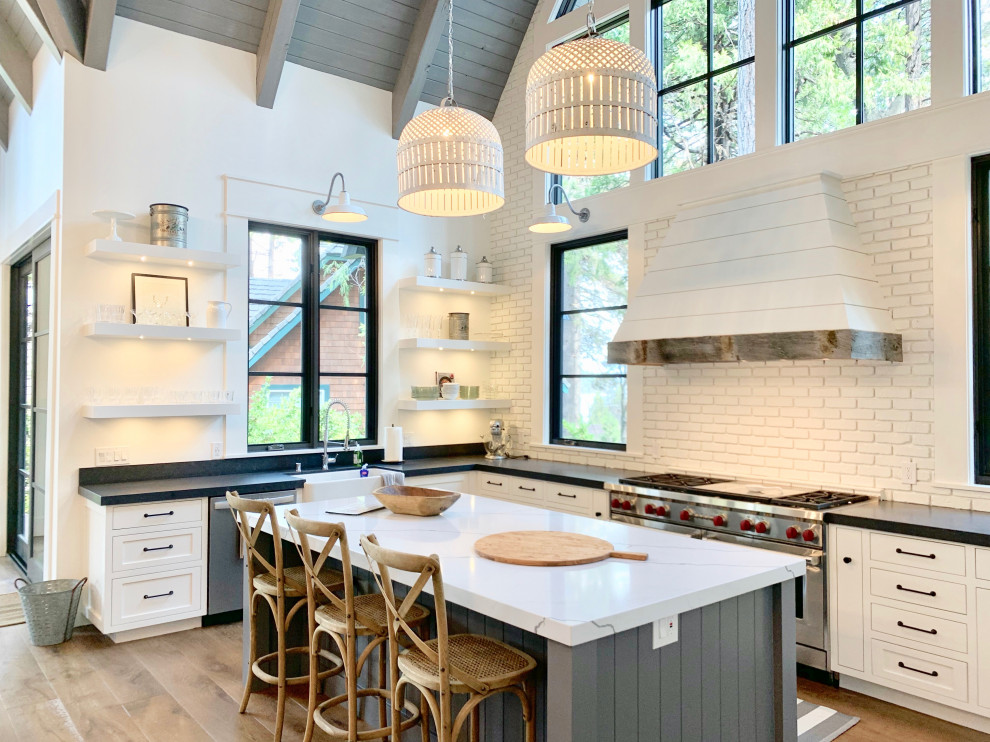 Design ideas for a farmhouse l-shaped kitchen in Los Angeles with a belfast sink, shaker cabinets, white cabinets, white splashback, brick splashback, stainless steel appliances, medium hardwood flooring, an island, brown floors and black worktops.