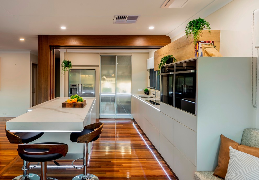 Große Moderne Wohnküche in L-Form mit flächenbündigen Schrankfronten, beigen Schränken, Quarzwerkstein-Arbeitsplatte, schwarzen Elektrogeräten, braunem Holzboden, Kücheninsel, braunem Boden und weißer Arbeitsplatte in Brisbane