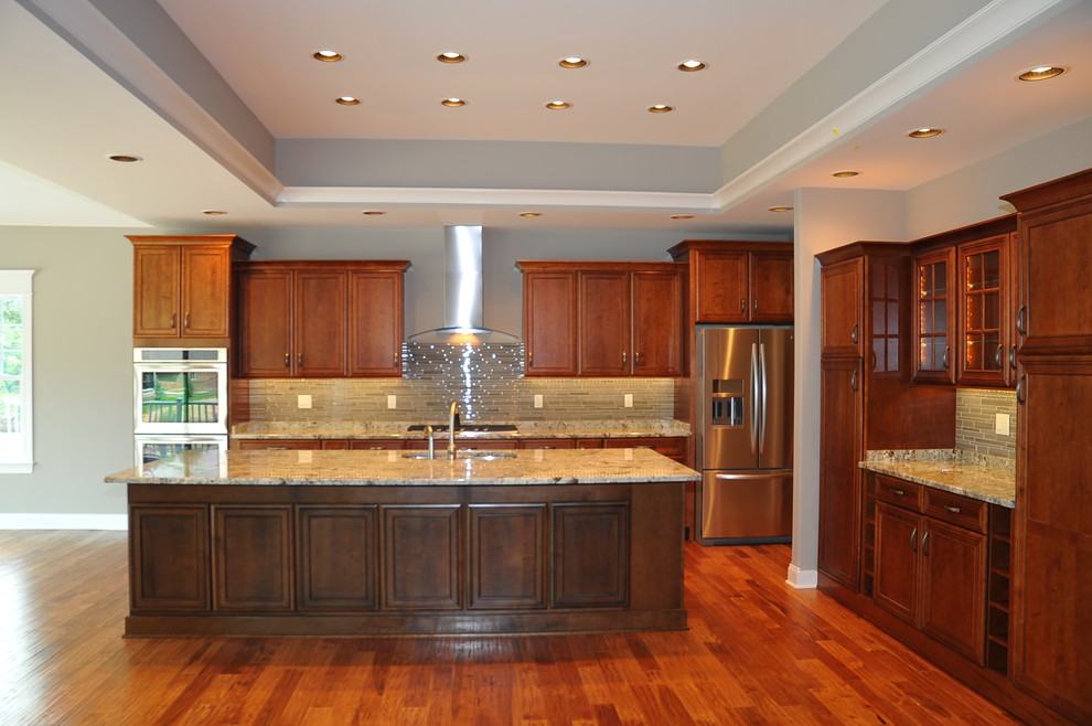 Amerikansk inredning av ett stort kök, med en undermonterad diskho, luckor med upphöjd panel, bruna skåp, granitbänkskiva, beige stänkskydd, stänkskydd i glaskakel, rostfria vitvaror, ljust trägolv och en köksö