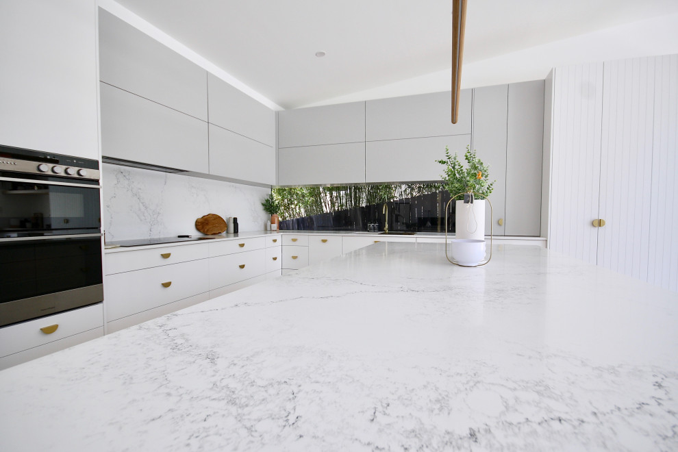 Example of a large minimalist l-shaped medium tone wood floor and brown floor open concept kitchen design in Sydney with an undermount sink, flat-panel cabinets, white cabinets, quartz countertops, white backsplash, stone slab backsplash, stainless steel appliances, an island and white countertops