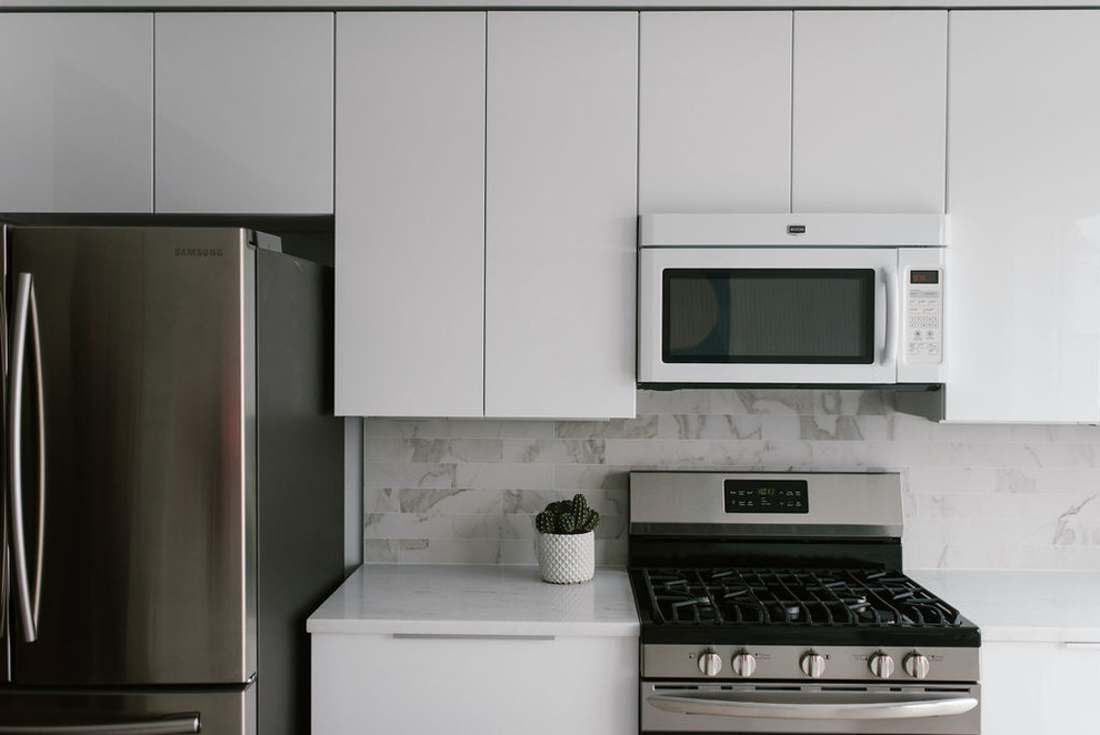 Photo of a small modern single-wall kitchen/diner in Baltimore with a double-bowl sink, flat-panel cabinets, white cabinets, marble worktops, white splashback, marble splashback, stainless steel appliances, light hardwood flooring, an island, beige floors and white worktops.