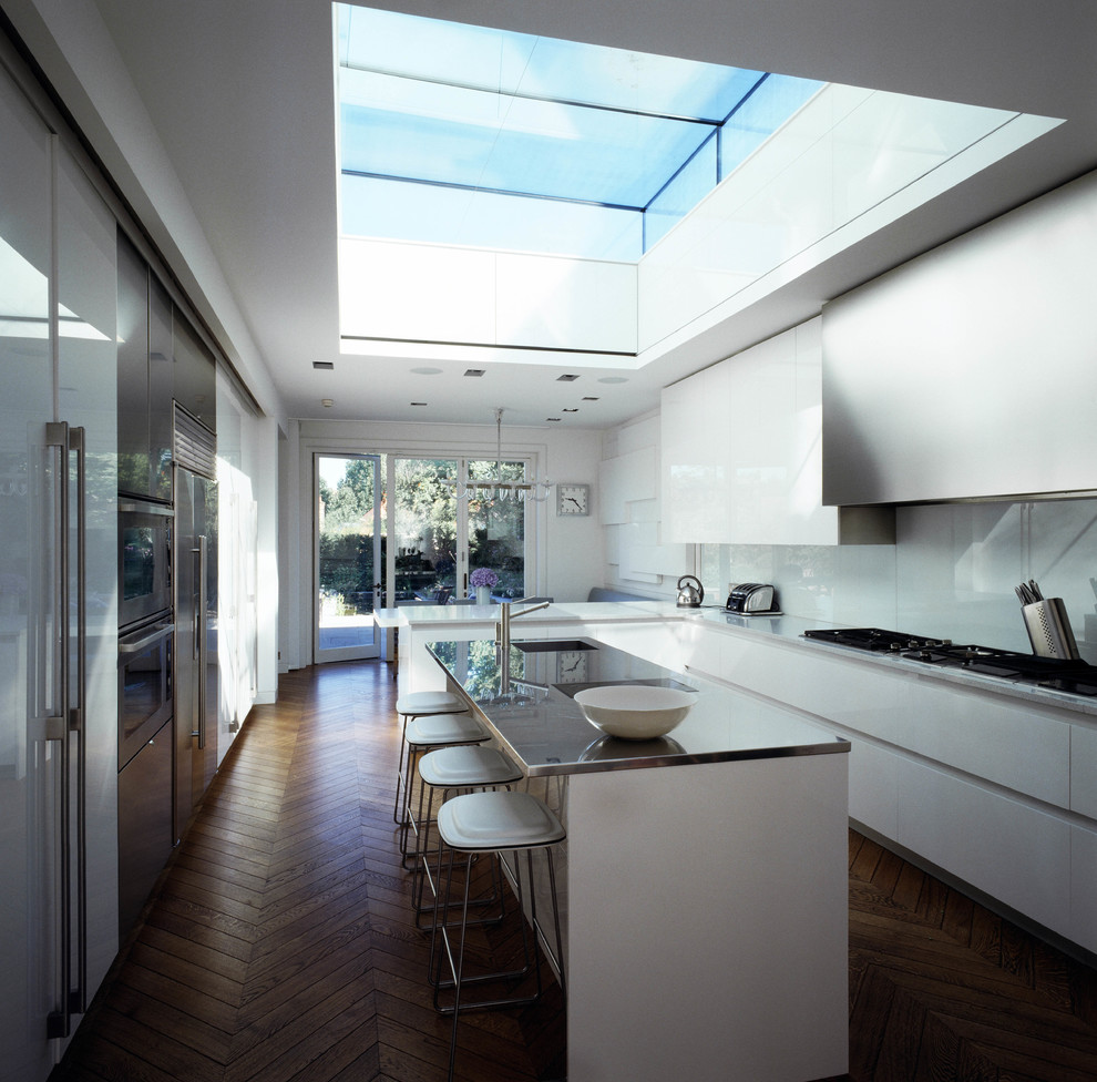 Cette photo montre une cuisine américaine parallèle tendance avec un placard à porte plane, une crédence blanche, un électroménager en acier inoxydable et plafond verrière.