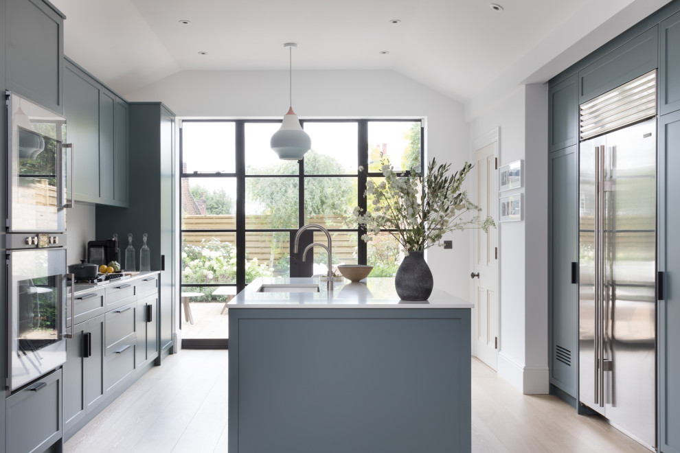Design ideas for a large contemporary galley kitchen in London with a submerged sink, shaker cabinets, blue cabinets, white splashback, stainless steel appliances, an island, beige floors and white worktops.