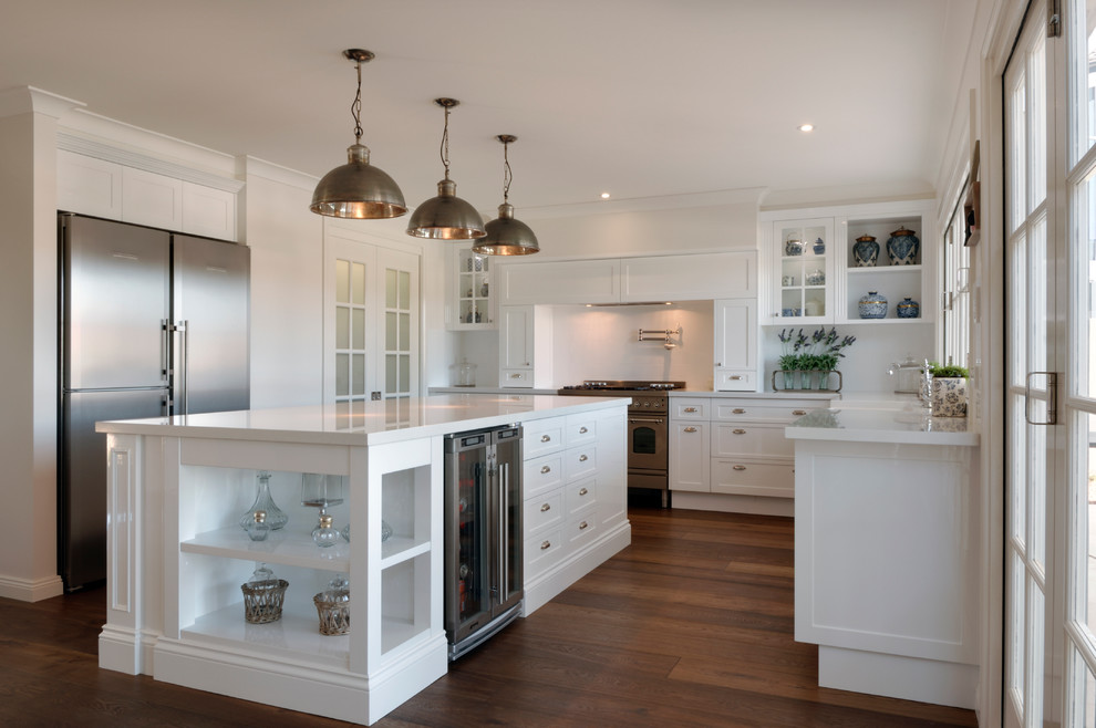 This is an example of a large traditional u-shaped kitchen in Brisbane with a belfast sink, shaker cabinets, white cabinets, engineered stone countertops, white splashback, metro tiled splashback, stainless steel appliances, medium hardwood flooring and an island.