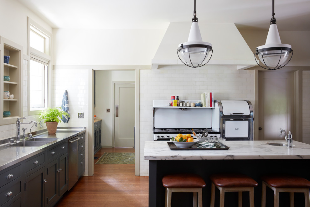 Mediterranean l-shaped kitchen in Los Angeles with a double-bowl sink, shaker cabinets, grey cabinets, stainless steel worktops, white splashback, metro tiled splashback, white appliances, medium hardwood flooring, an island, brown floors and grey worktops.