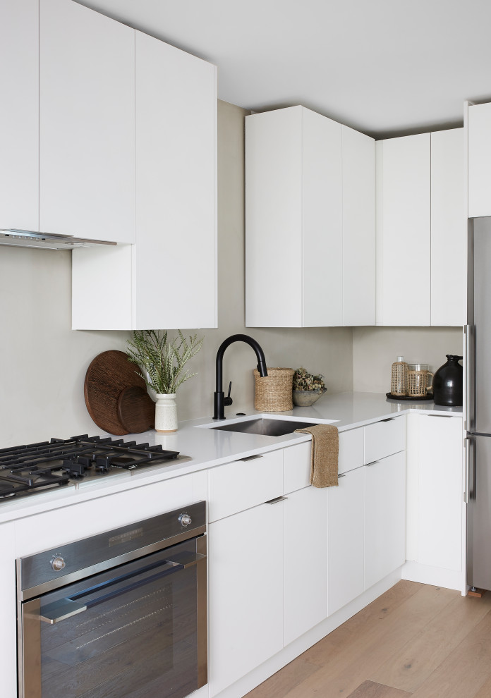 Modern inredning av ett litet vit vitt l-kök, med släta luckor, vita skåp, bänkskiva i koppar, grått stänkskydd, rostfria vitvaror, brunt golv, en undermonterad diskho och mellanmörkt trägolv