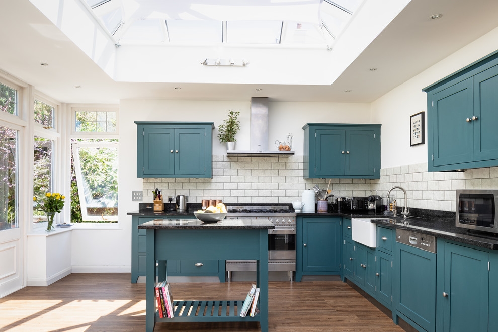 Klassische Küche in L-Form mit Landhausspüle, Schrankfronten im Shaker-Stil, Küchenrückwand in Grau, Rückwand aus Metrofliesen, Küchengeräten aus Edelstahl, Kücheninsel, schwarzer Arbeitsplatte, blauen Schränken, Granit-Arbeitsplatte und dunklem Holzboden in Surrey