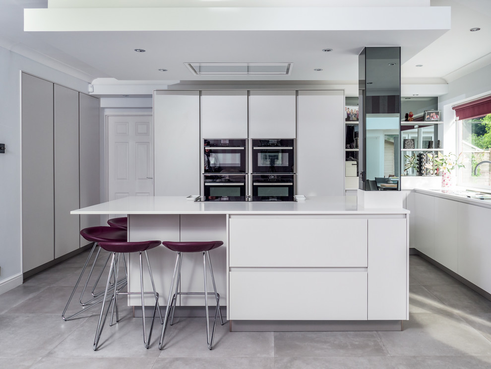 Contemporary l-shaped kitchen in Berkshire with flat-panel cabinets, white cabinets, window splashback, black appliances, an island, grey floors and white worktops.