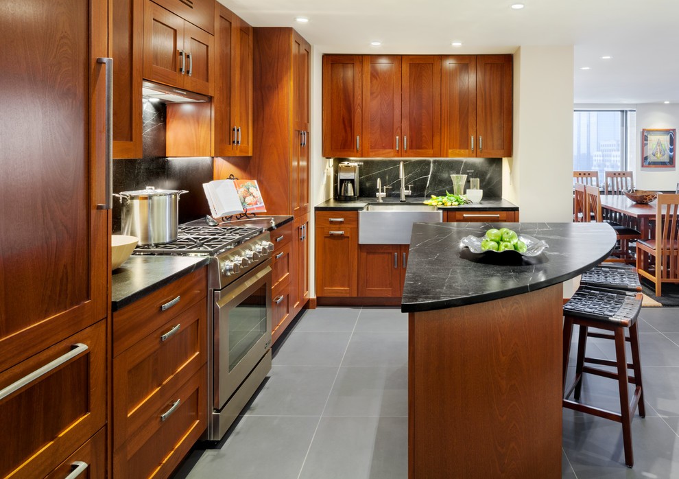 Example of an arts and crafts l-shaped limestone floor open concept kitchen design in Boston with a farmhouse sink, shaker cabinets, medium tone wood cabinets, soapstone countertops, black backsplash, stone slab backsplash, stainless steel appliances and an island