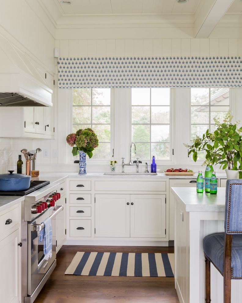 Inredning av ett maritimt vit vitt kök, med en rustik diskho, vita skåp, marmorbänkskiva, vitt stänkskydd, rostfria vitvaror, mellanmörkt trägolv, en köksö och brunt golv