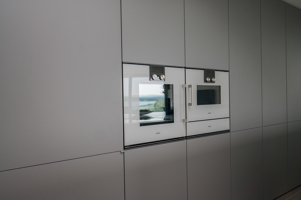This is an example of a large contemporary single-wall open plan kitchen in Dorset with a built-in sink, flat-panel cabinets, grey cabinets, composite countertops, white appliances, porcelain flooring and an island.