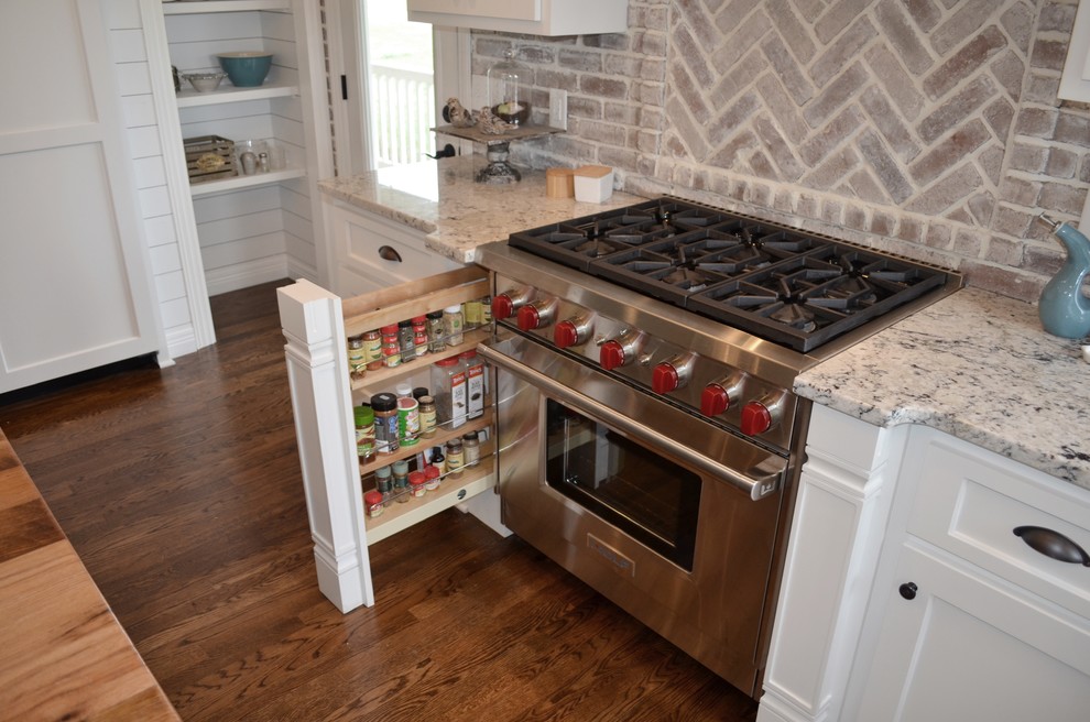 Inspiration for a mid-sized cottage u-shaped medium tone wood floor and brown floor enclosed kitchen remodel in Nashville with a farmhouse sink, shaker cabinets, white cabinets, granite countertops, beige backsplash, brick backsplash, paneled appliances and an island