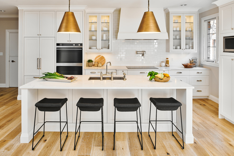 Example of a mid-sized transitional light wood floor and beige floor kitchen design in Vancouver with white cabinets, quartz countertops, white backsplash, stainless steel appliances, an island, white countertops, an undermount sink, glass-front cabinets and subway tile backsplash