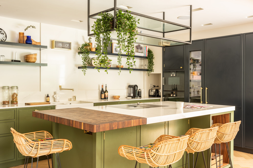 Beach style kitchen in London with a belfast sink, flat-panel cabinets, green cabinets, white splashback, integrated appliances, light hardwood flooring, an island and white worktops.