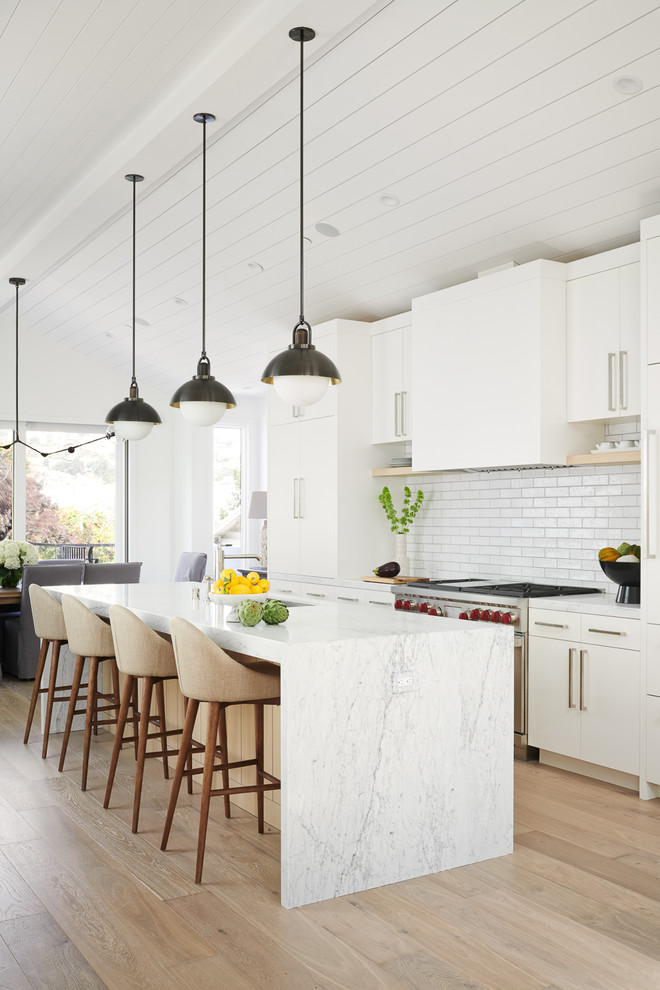 Ejemplo de cocina comedor tradicional renovada, grande, con fregadero bajoencimera, armarios con paneles lisos, puertas de armario blancas, encimera de mármol, salpicadero blanco, salpicadero de azulejos de cerámica, electrodomésticos con paneles, suelo de madera clara, una isla, encimeras blancas y suelo marrón
