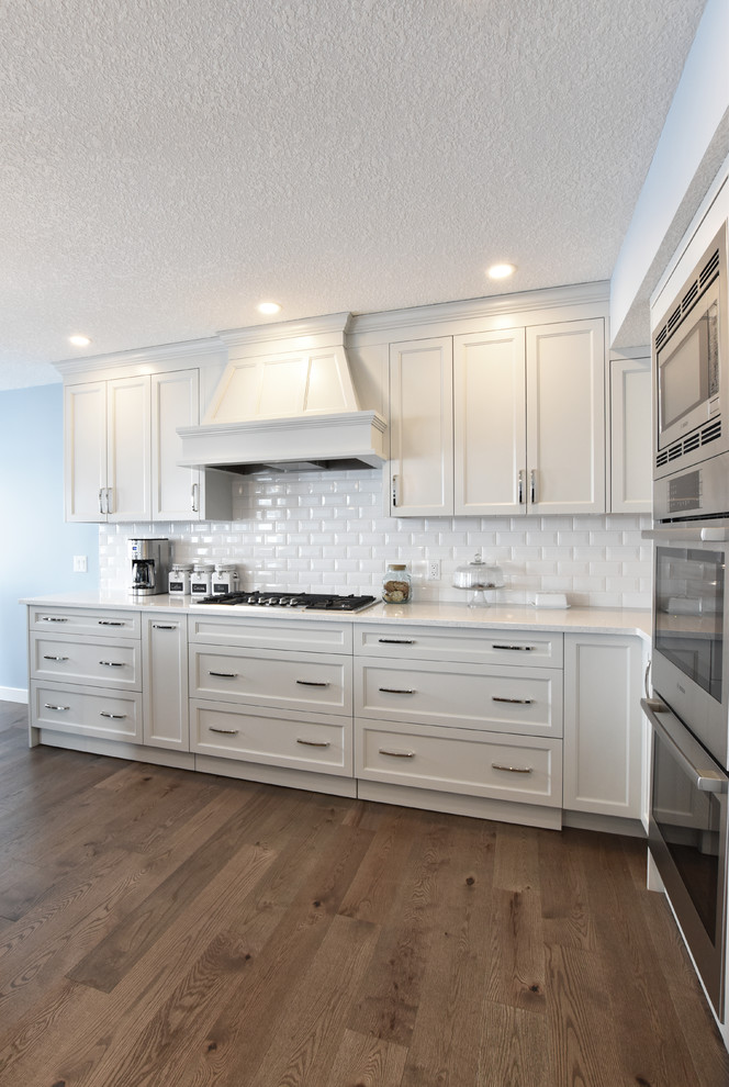 Photo of a large traditional l-shaped open plan kitchen in Calgary with a submerged sink, recessed-panel cabinets, grey cabinets, quartz worktops, white splashback, ceramic splashback, stainless steel appliances, medium hardwood flooring, an island and brown floors.