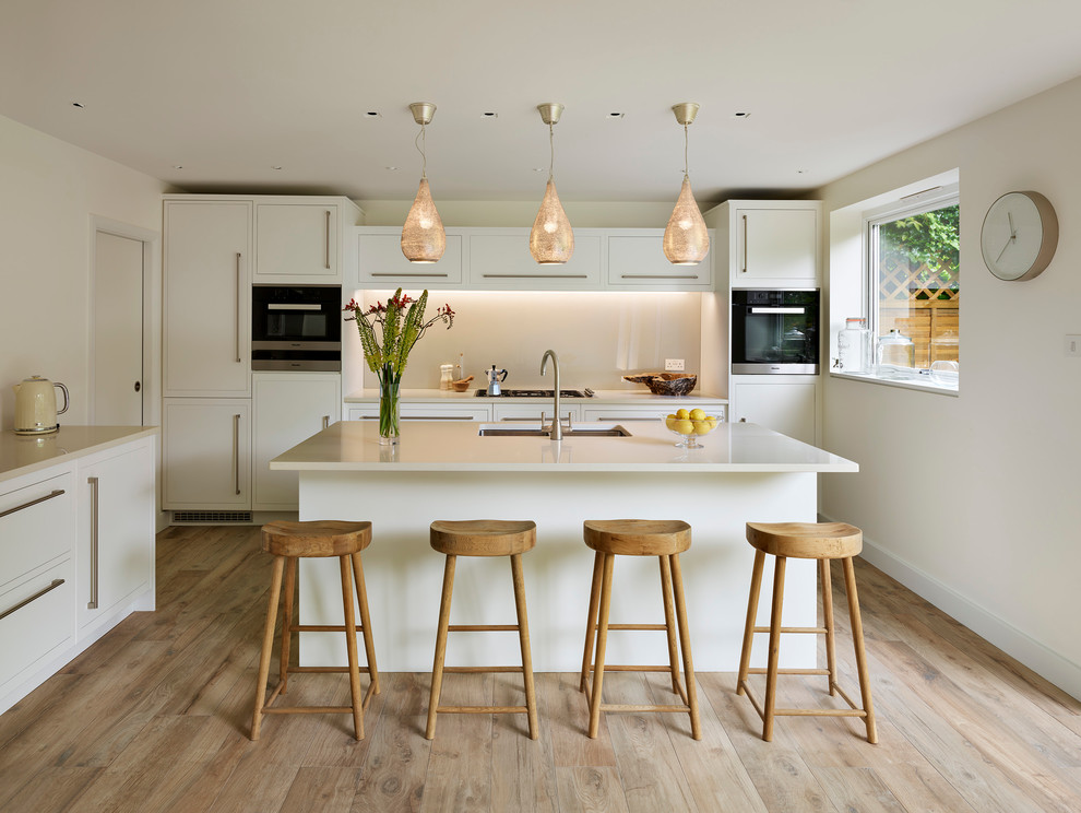 Modern inredning av ett l-kök, med en undermonterad diskho, släta luckor, vita skåp, beige stänkskydd, glaspanel som stänkskydd, svarta vitvaror, ljust trägolv och en köksö