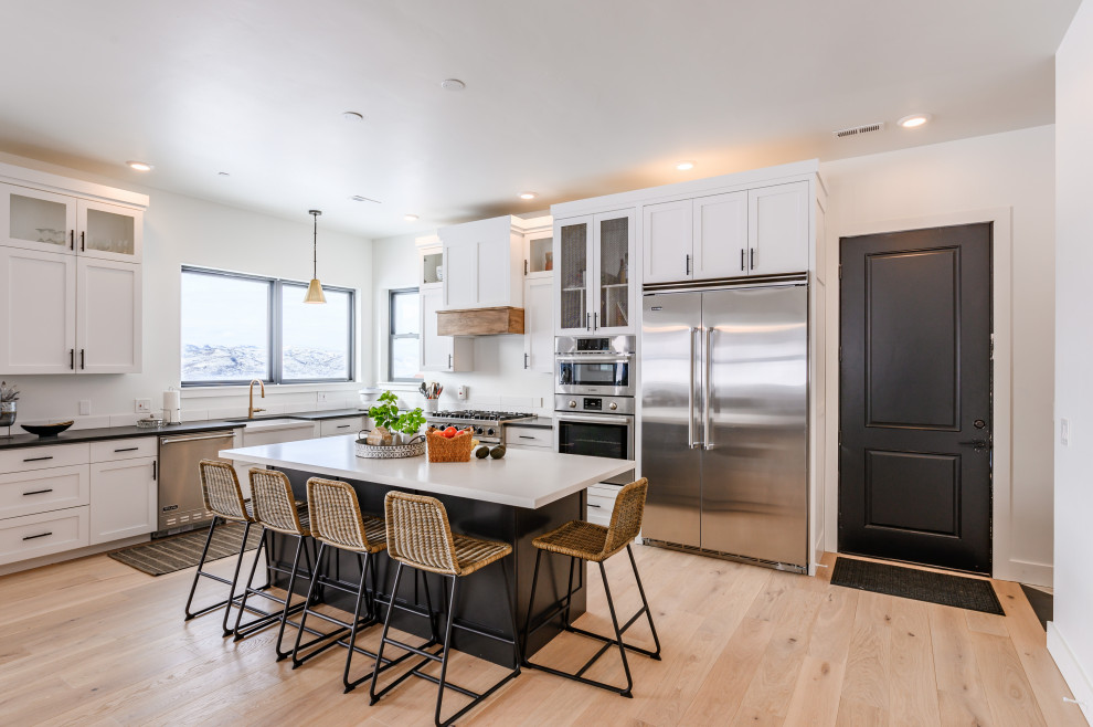 This is an example of a country l-shaped open plan kitchen in Salt Lake City with a belfast sink, shaker cabinets, white cabinets, white splashback, stainless steel appliances, medium hardwood flooring, an island, brown floors and white worktops.