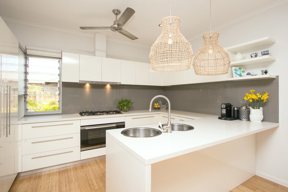 This is an example of a medium sized beach style u-shaped kitchen/diner in Dunedin with a double-bowl sink, flat-panel cabinets, white cabinets, engineered stone countertops, grey splashback, porcelain splashback, stainless steel appliances and bamboo flooring.