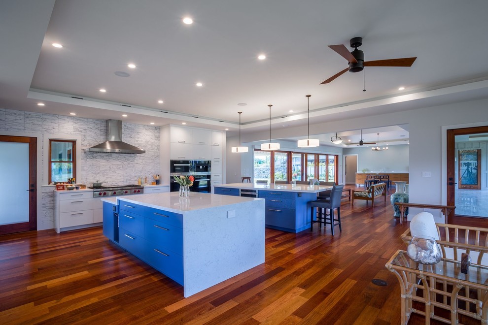 Inspiration for a medium sized traditional single-wall open plan kitchen in Hawaii with flat-panel cabinets, white cabinets, engineered stone countertops, white splashback, stone tiled splashback, stainless steel appliances, medium hardwood flooring, multiple islands and brown floors.