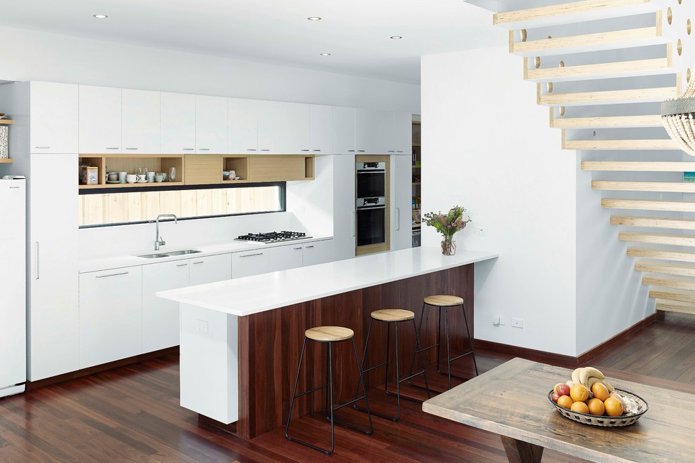 Modern inredning av ett mellanstort vit vitt kök, med en undermonterad diskho, vita skåp, bänkskiva i kvarts, vitt stänkskydd, rostfria vitvaror, mörkt trägolv, en köksö, släta luckor och brunt golv