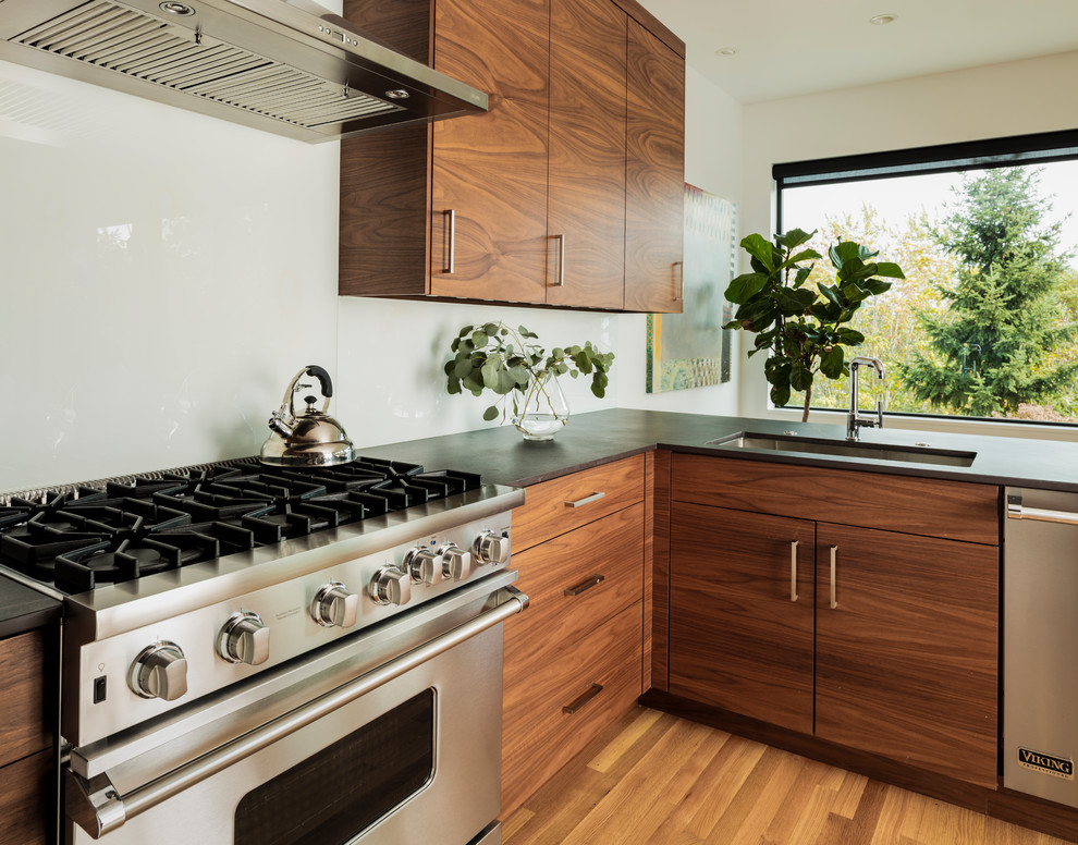 Foto på ett retro svart kök, med en undermonterad diskho, släta luckor, skåp i mellenmörkt trä, bänkskiva i kvarts, vitt stänkskydd, glaspanel som stänkskydd, rostfria vitvaror, ljust trägolv, en halv köksö och brunt golv