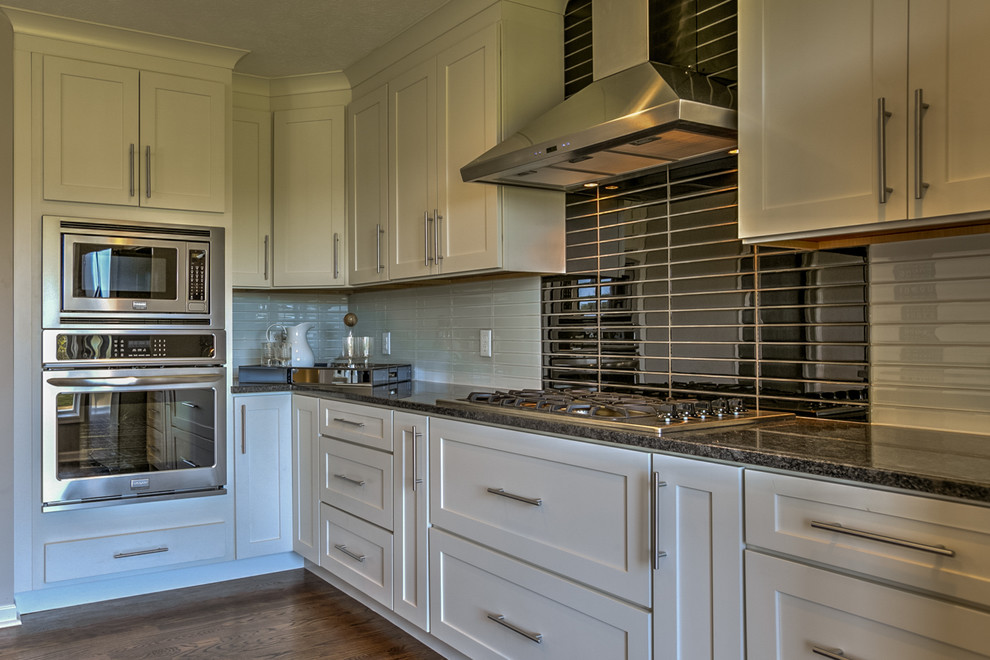 Classic l-shaped kitchen pantry in Omaha with a submerged sink, shaker cabinets, white cabinets, granite worktops, black splashback, glass tiled splashback, stainless steel appliances, medium hardwood flooring and an island.