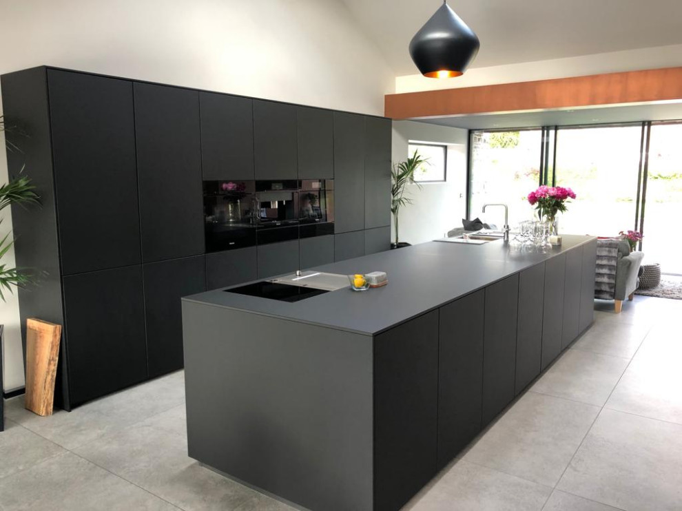 Modern inredning av ett stort svart linjärt svart kök med öppen planlösning, med en dubbel diskho, släta luckor, svarta skåp, bänkskiva i koppar, svarta vitvaror, klinkergolv i porslin, en köksö och beiget golv