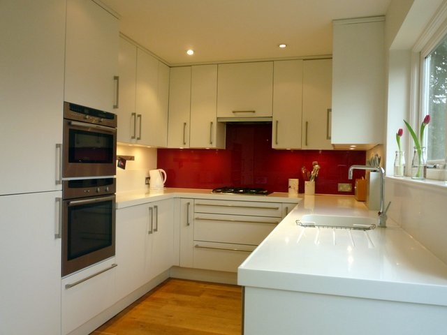 Photo of a medium sized modern u-shaped kitchen/diner in London with an integrated sink, flat-panel cabinets, white cabinets, composite countertops, red splashback, glass sheet splashback, stainless steel appliances, medium hardwood flooring and no island.