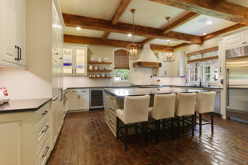 Foto på ett stort lantligt kök, med en undermonterad diskho, luckor med profilerade fronter, vita skåp, granitbänkskiva, vitt stänkskydd, rostfria vitvaror, tegelgolv, en köksö och stänkskydd i tunnelbanekakel
