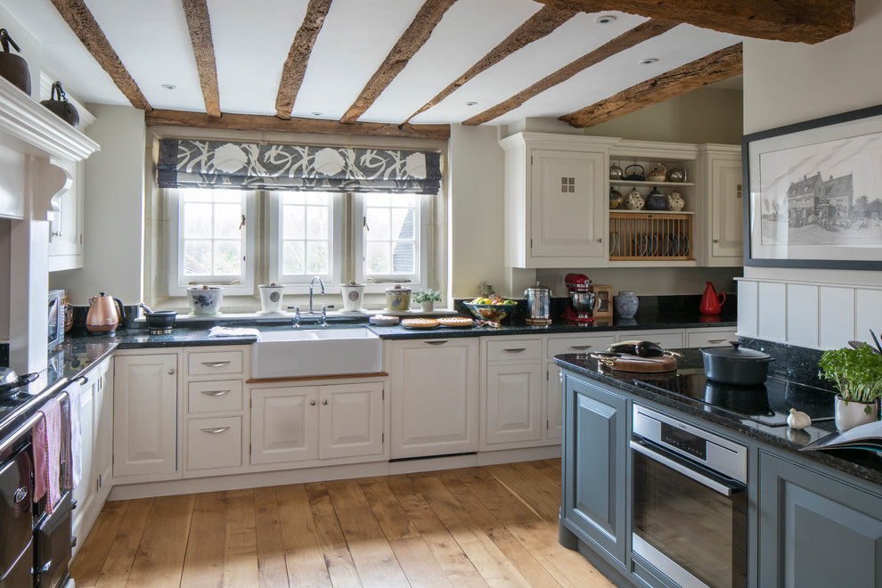 Inspiration for a medium sized farmhouse l-shaped kitchen in Other with a belfast sink, raised-panel cabinets, stainless steel appliances, light hardwood flooring, beige floors and black worktops.