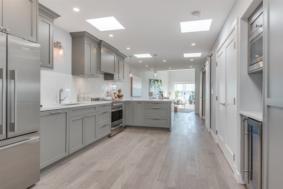 Example of a mid-sized beach style l-shaped light wood floor and gray floor open concept kitchen design in Vancouver with an undermount sink, shaker cabinets, gray cabinets, quartz countertops, white backsplash, subway tile backsplash, stainless steel appliances, a peninsula and white countertops