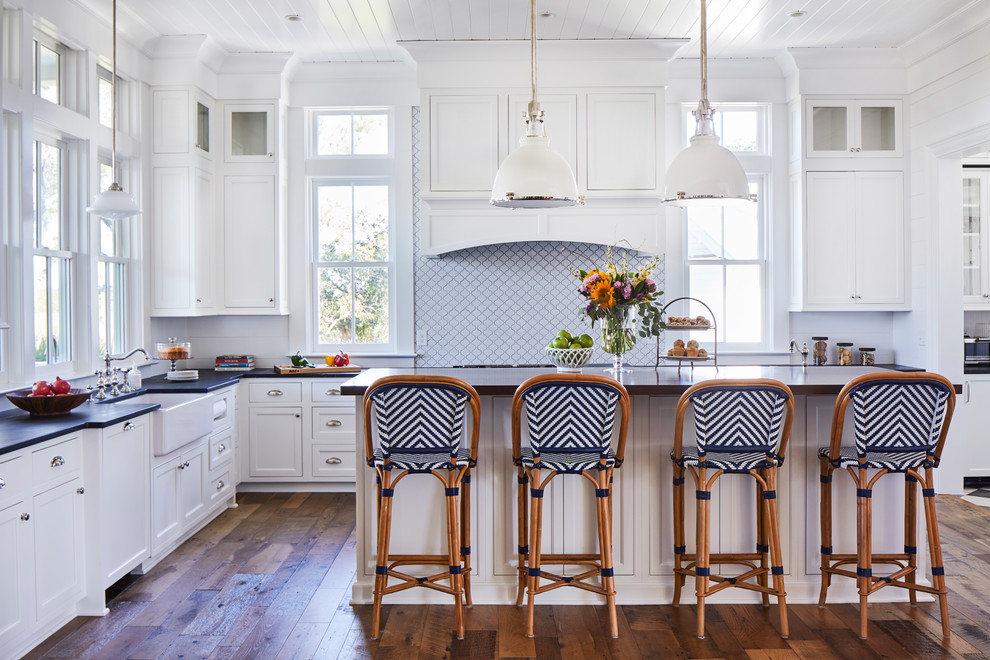 Inspiration for a large coastal l-shaped medium tone wood floor and brown floor kitchen remodel in Charleston with a farmhouse sink, soapstone countertops, white backsplash, porcelain backsplash, an island, black countertops, white cabinets and shaker cabinets