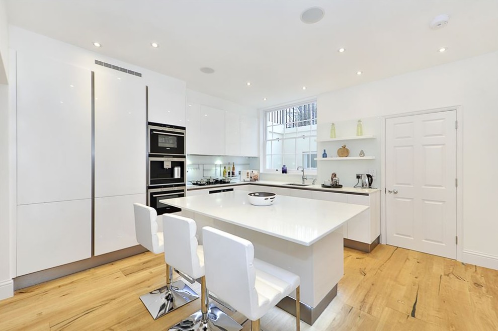 Große Moderne Küche in L-Form mit flächenbündigen Schrankfronten, weißen Schränken, Glasrückwand, Kücheninsel, weißer Arbeitsplatte, Unterbauwaschbecken, Elektrogeräten mit Frontblende, hellem Holzboden und beigem Boden in London