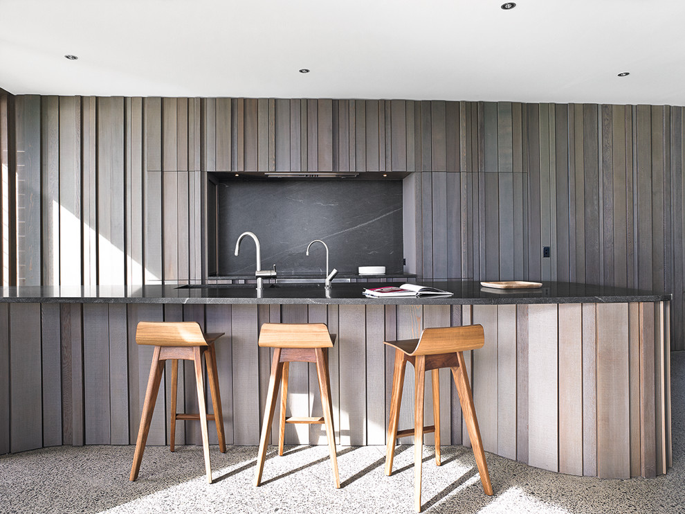 Modern inredning av ett kök, med skåp i mellenmörkt trä, granitbänkskiva, svart stänkskydd, rostfria vitvaror, en undermonterad diskho, luckor med profilerade fronter, terrazzogolv, en köksö och grått golv